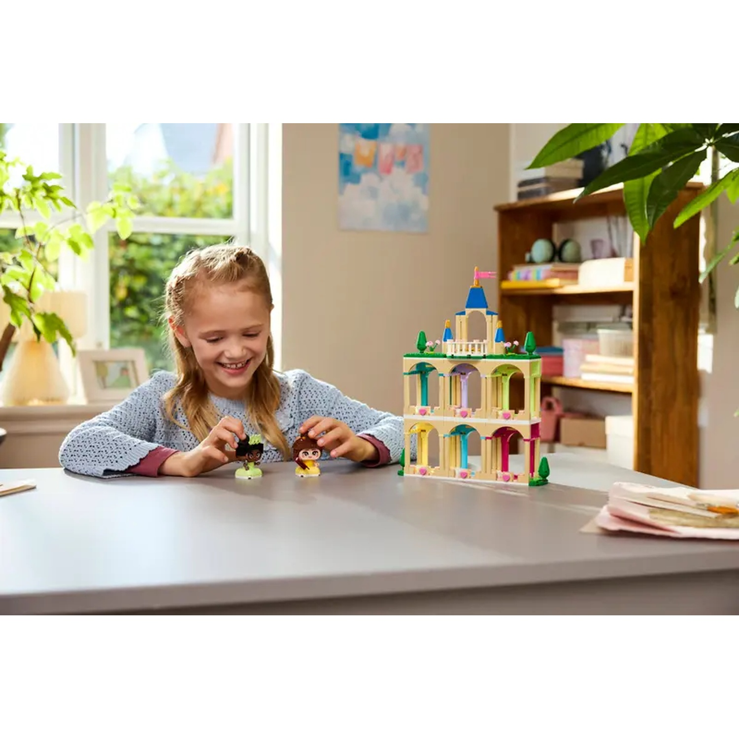 Een meisje glimlacht, spelend met Lego-figuren en een kasteel. De scène is binnen, met een tafel, raam en boekenplank in beeld.