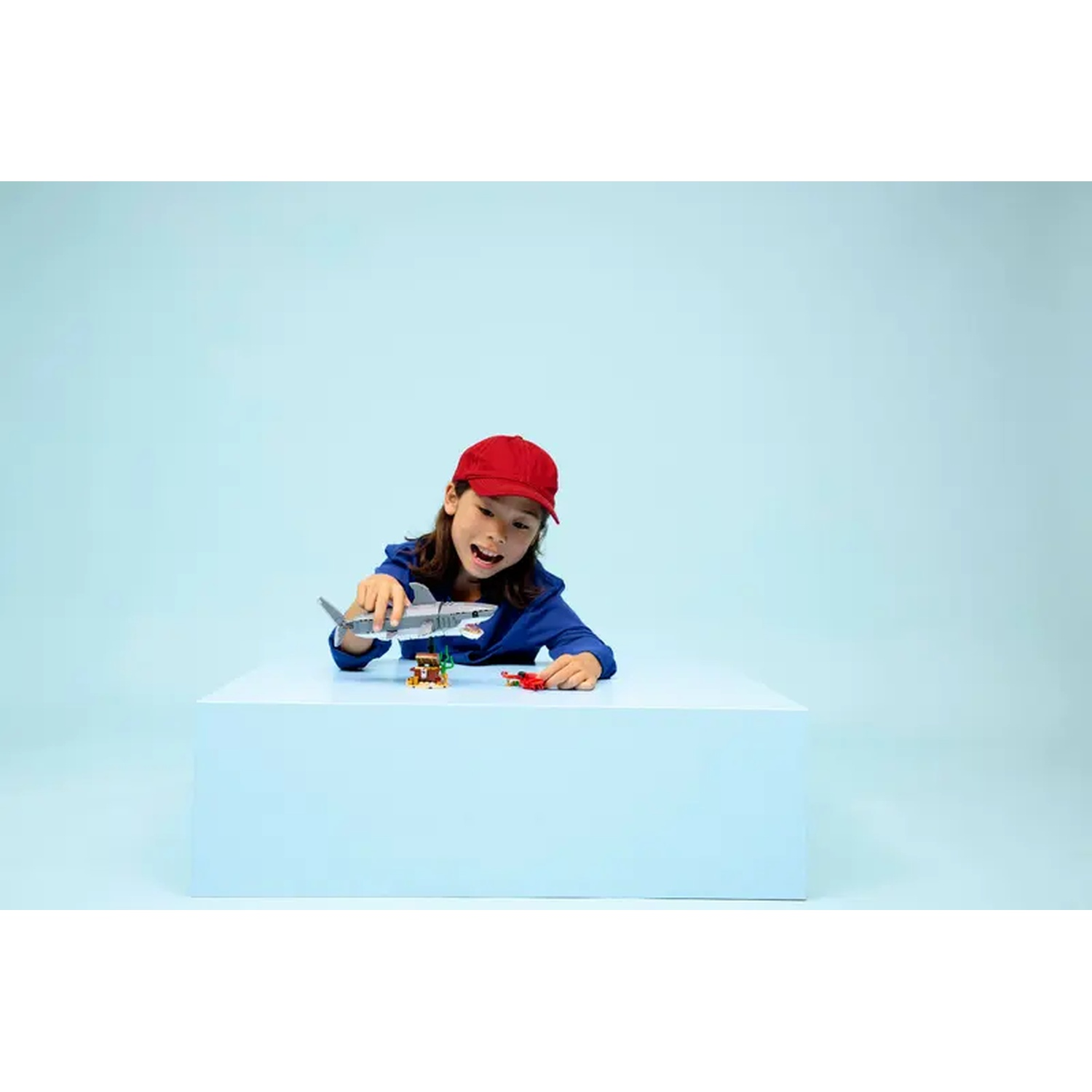 Een kind met een rode pet speelt met speelgoed, waaronder een haai, op een wit oppervlak tegen een blauwe achtergrond.