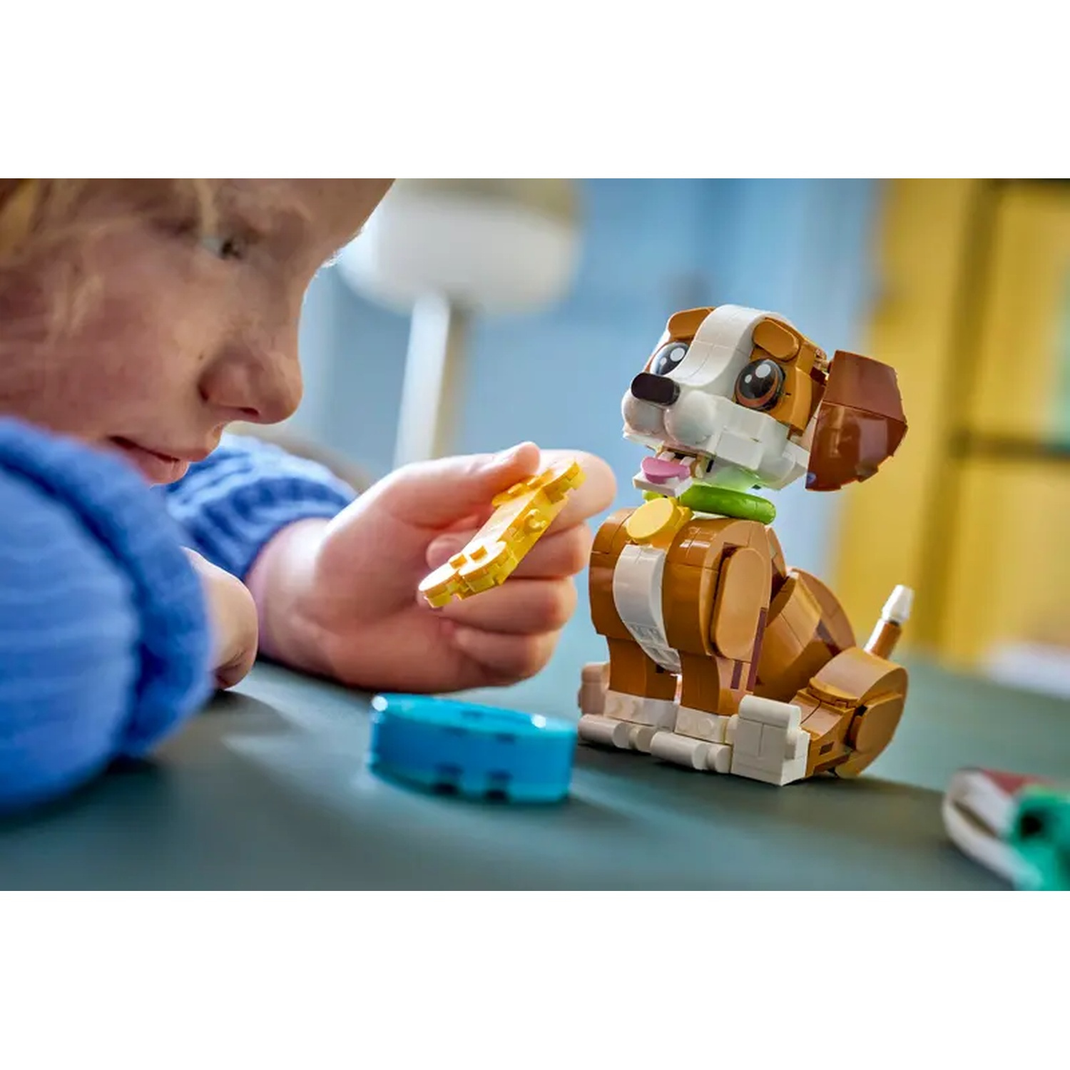 Een kind speelt met een Lego hond. Het kind houdt een geel Lego stuk vast, de hond zit op een tafel met andere Lego's.