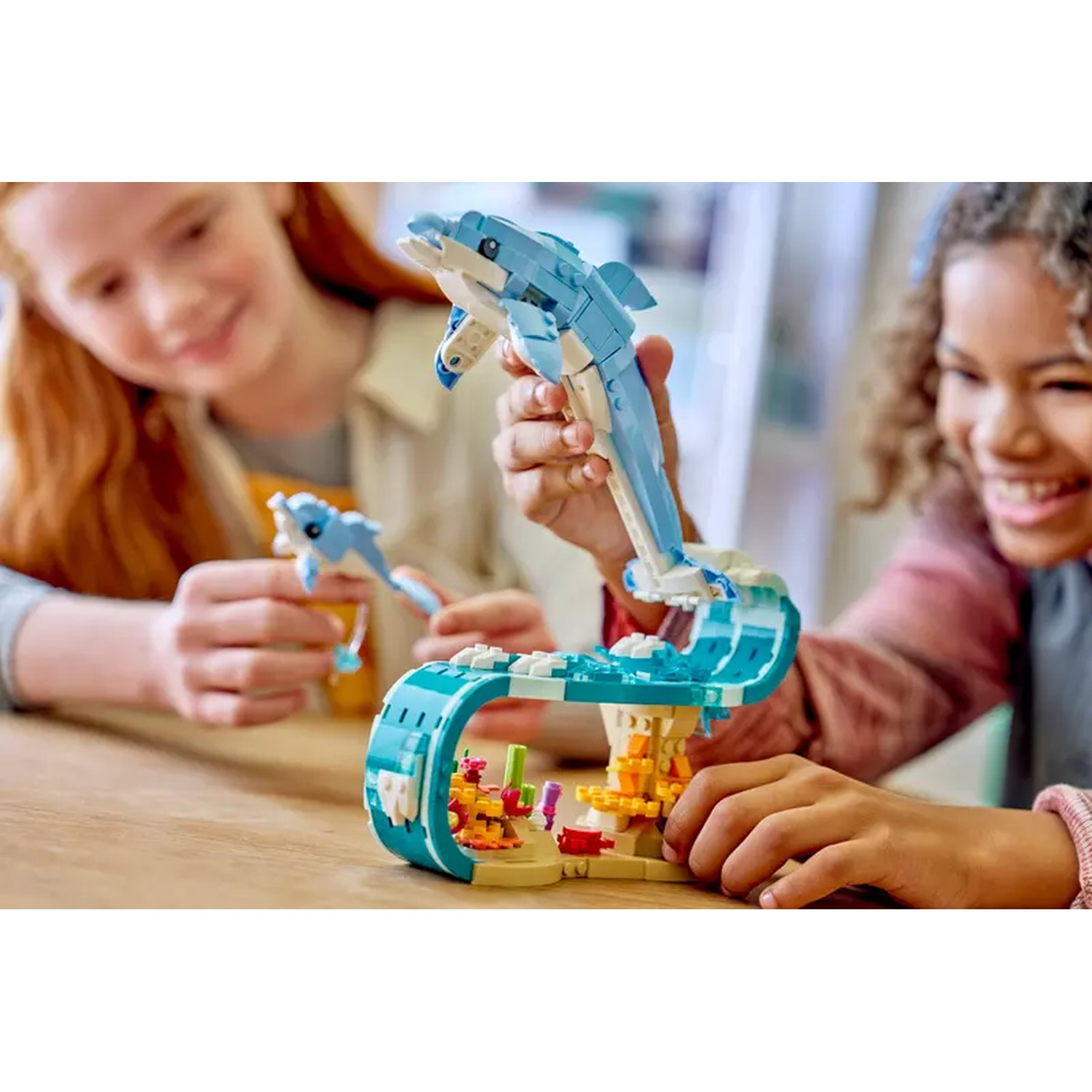 Deux filles jouent avec un ensemble de dauphins Lego. L'une tient un grand dauphin sautant au-dessus d'une vague, l'autre un petit dauphin.