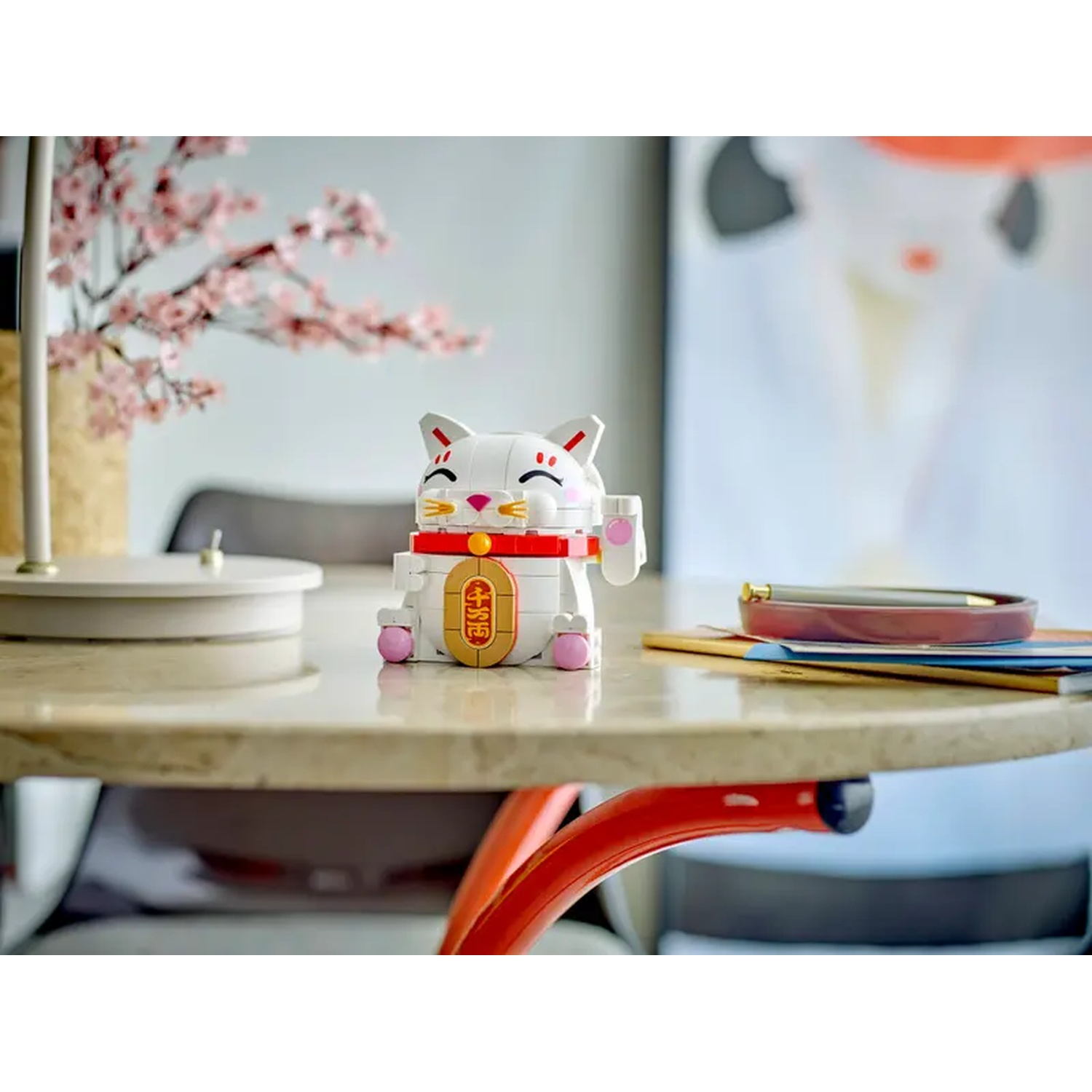 Een wit gelukskatfiguur op een tafel. Kersenbloesems op de achtergrond. Een pen en notitieboekjes staan ook op de tafel.