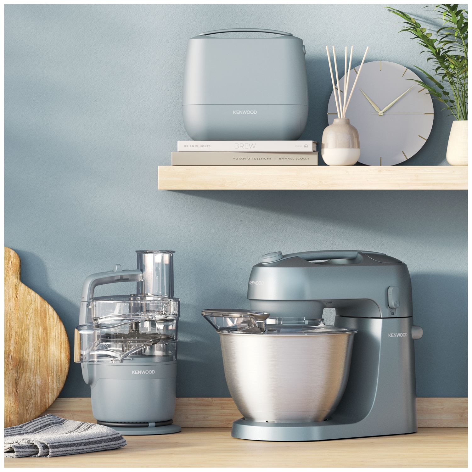 Kitchen appliances on wooden counter. Includes food processor, stand mixer, and other items. Shelf with clock and books in the background.