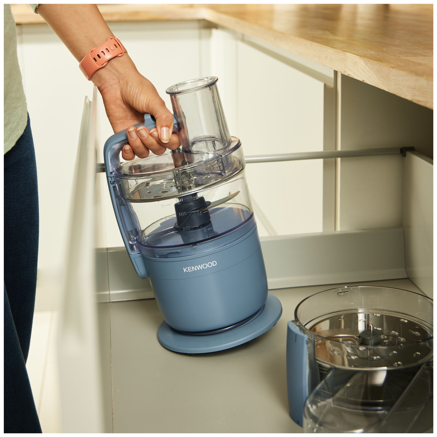 A person is holding a blue food processor. The processor is being placed in a white kitchen cabinet. A second part is near.
