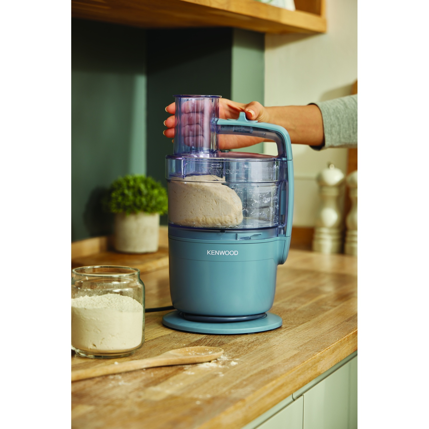A Kenwood food processor with dough inside, a hand is holding the lid. Flour in a jar and a wooden spoon are on the wooden counter.