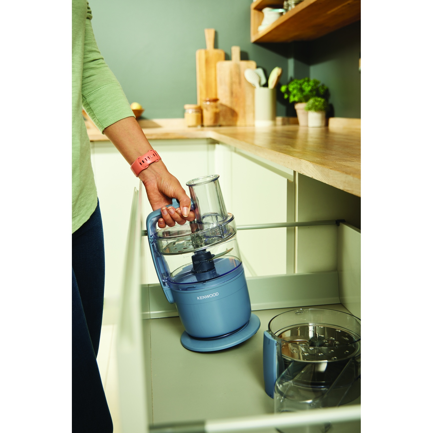 A person holds a Kenwood food processor. It's blue with clear attachments, placed in an open kitchen drawer. Wooden shelves are in the background.