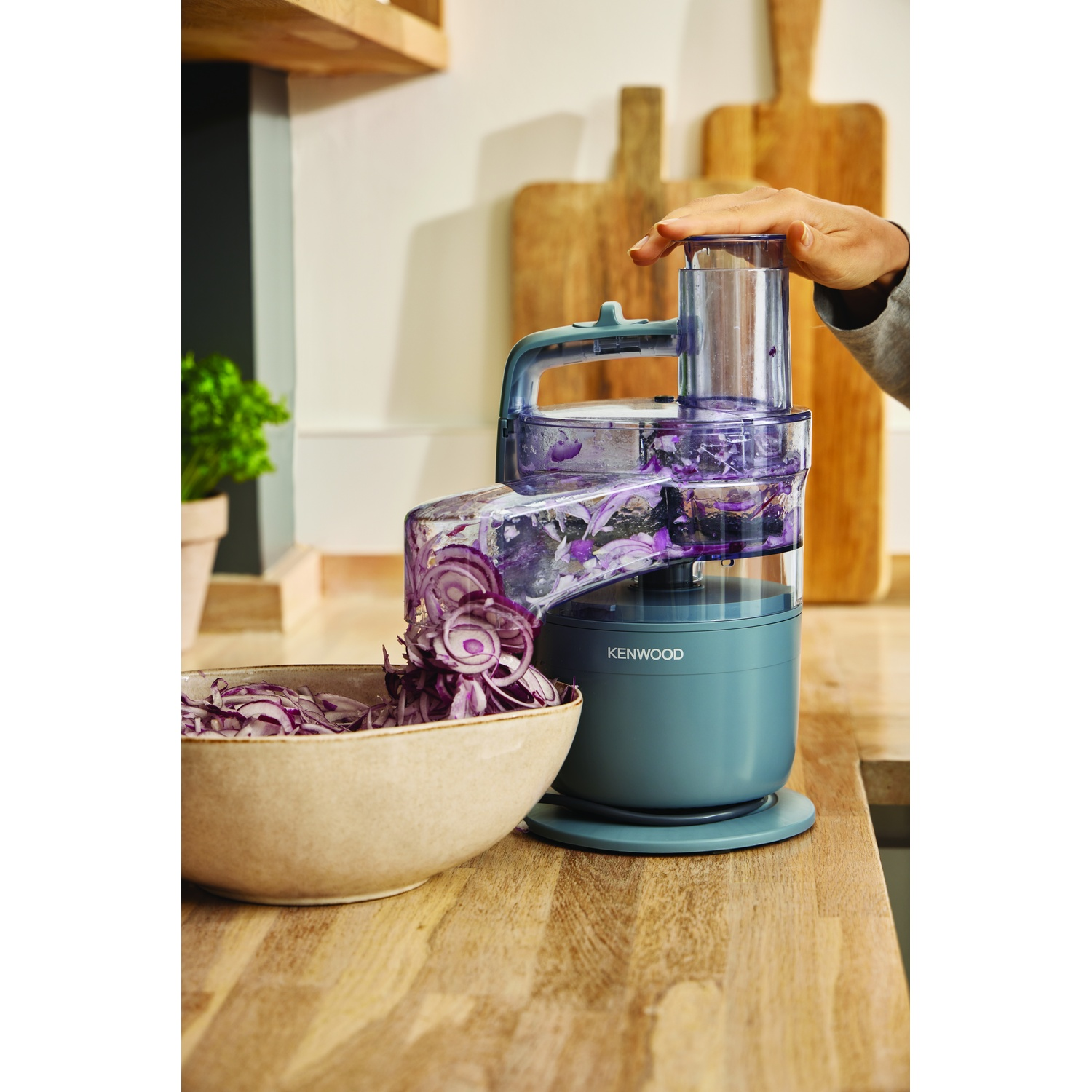 A food processor is slicing red onions. Sliced onions fall into a bowl. A hand is on the processor lid. Wooden boards in the background.