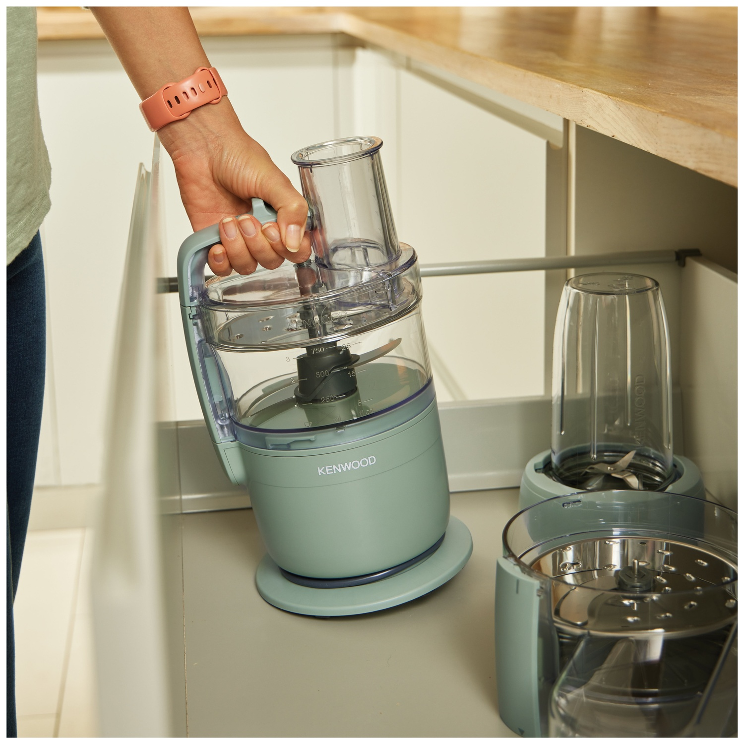 A person holding a light green Kenwood food processor. Other parts of the appliance are visible. The person is in a kitchen.