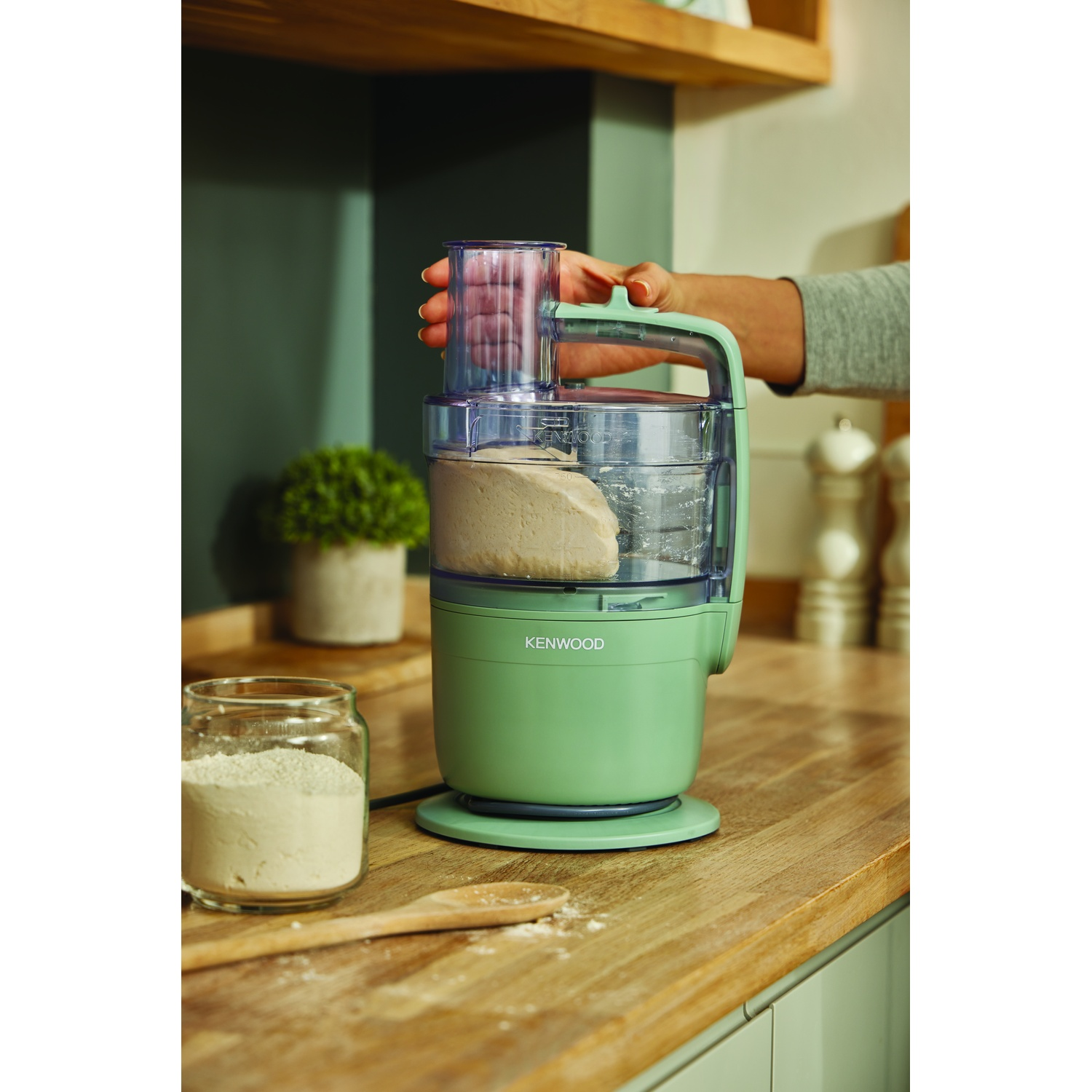 A green food processor with dough inside is held by a hand. Jar of flour and a wooden spoon are on the wooden counter.