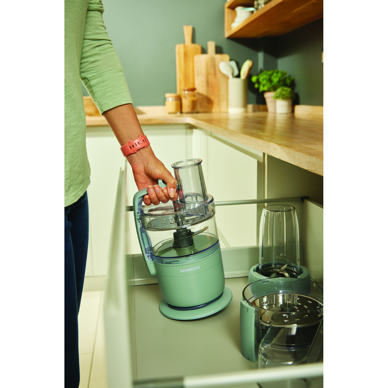 A person puts a green food processor in a drawer. Other parts and a blender are also in the drawer. Kitchen in the background.