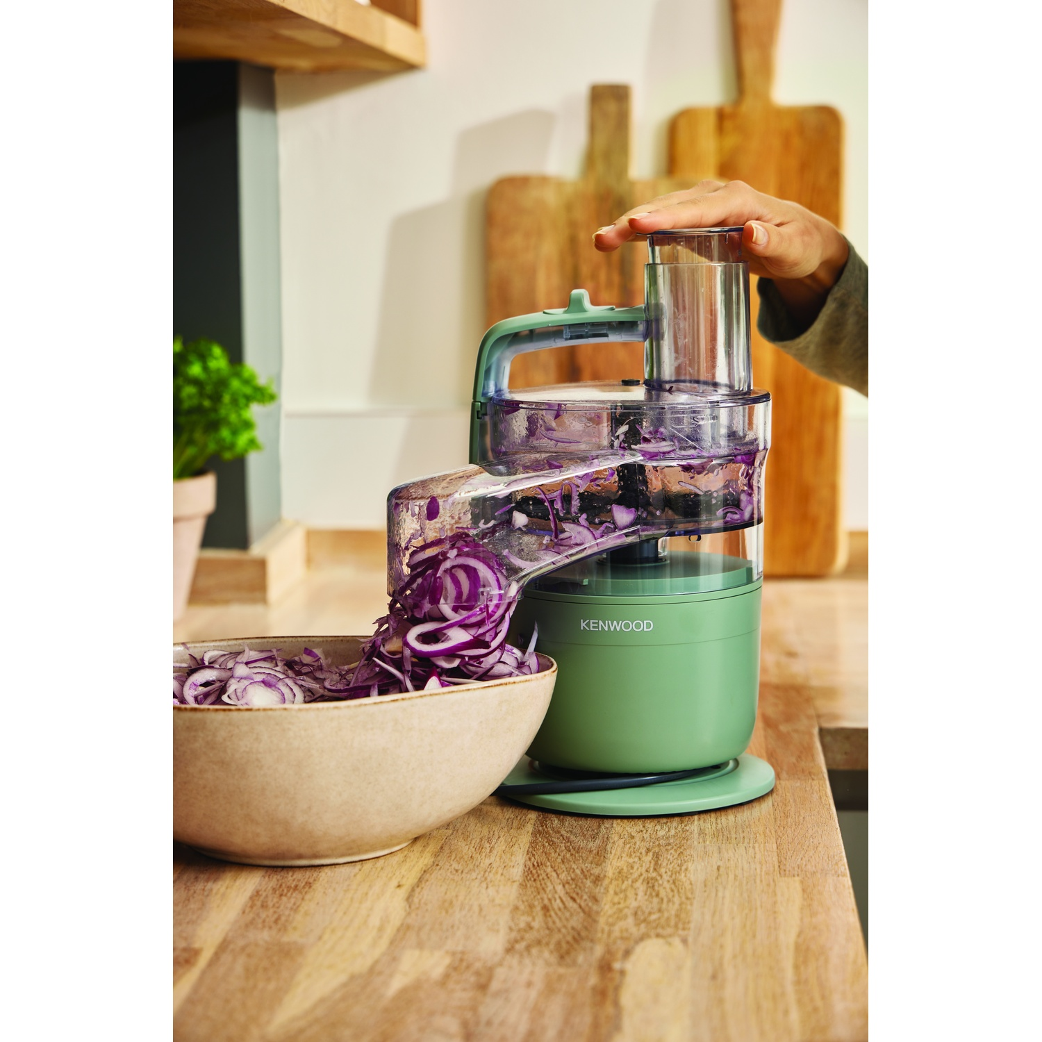 A green Kenwood food processor is slicing red onions into a bowl. A hand is holding the lid. Wooden boards are in the background.