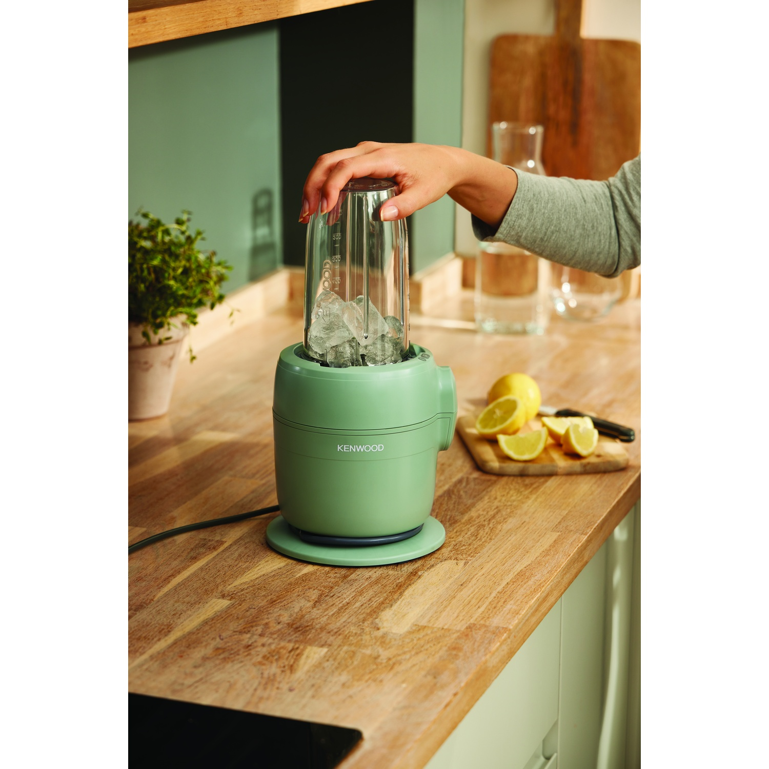 A hand places a glass on a green blender with ice inside. Sliced lemons and a knife sit on a cutting board. Wooden counter.