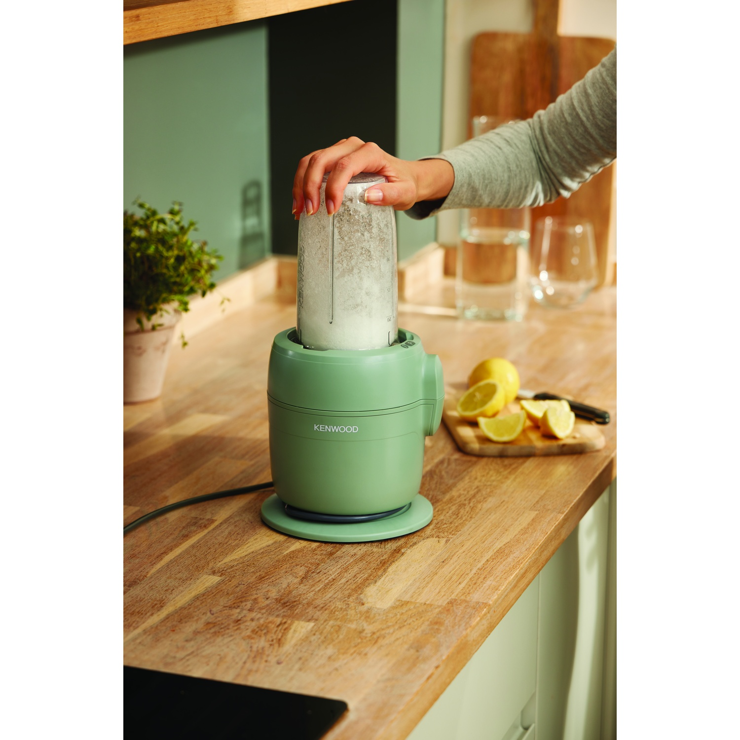 A person is using a green Kenwood blender. Lemons and a cutting board are on the wooden counter. A plant is in the background.
