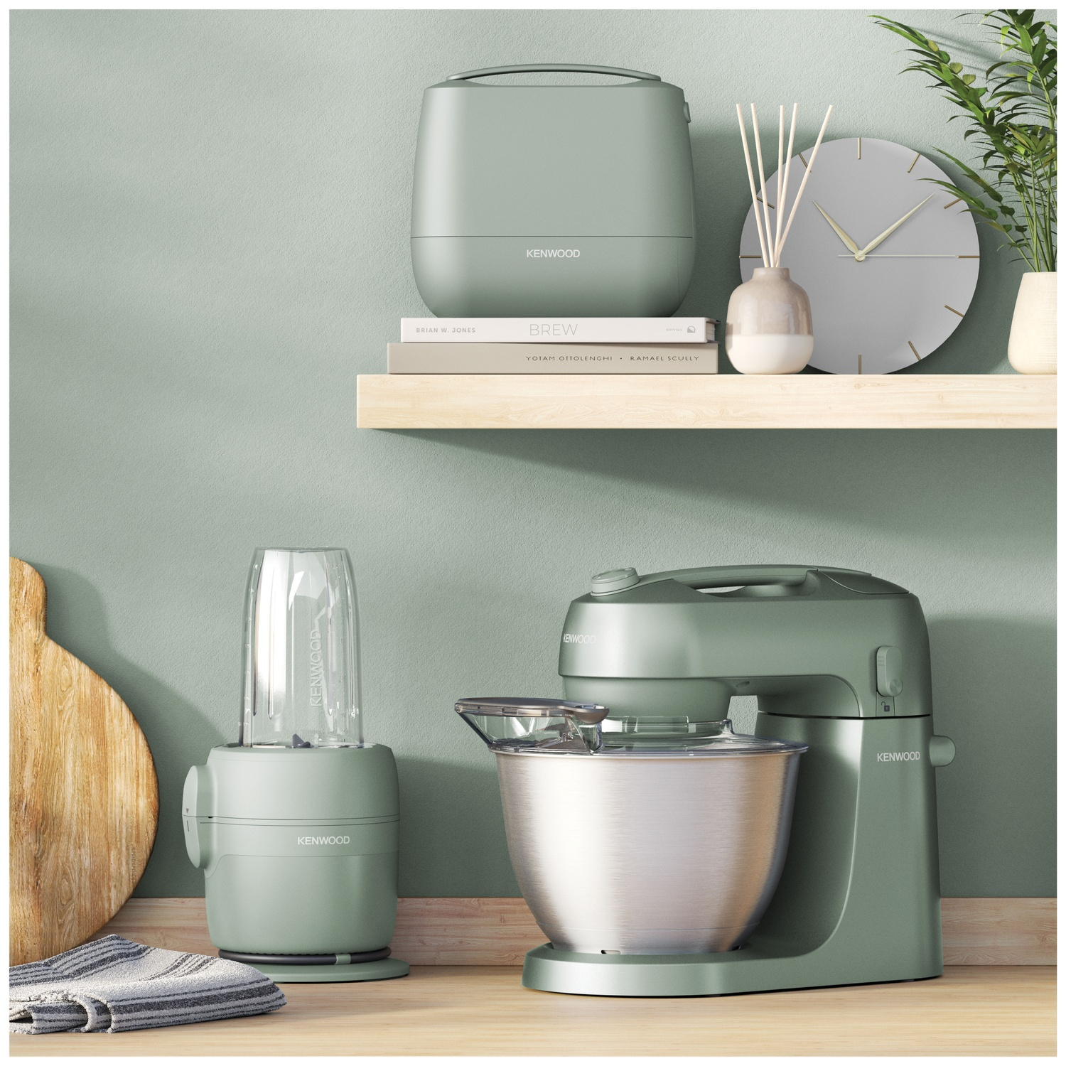 Scène de cuisine avec appareils Kenwood : mixeur, mélangeur et un petit appareil. Une horloge, des livres et des plantes sur une étagère.