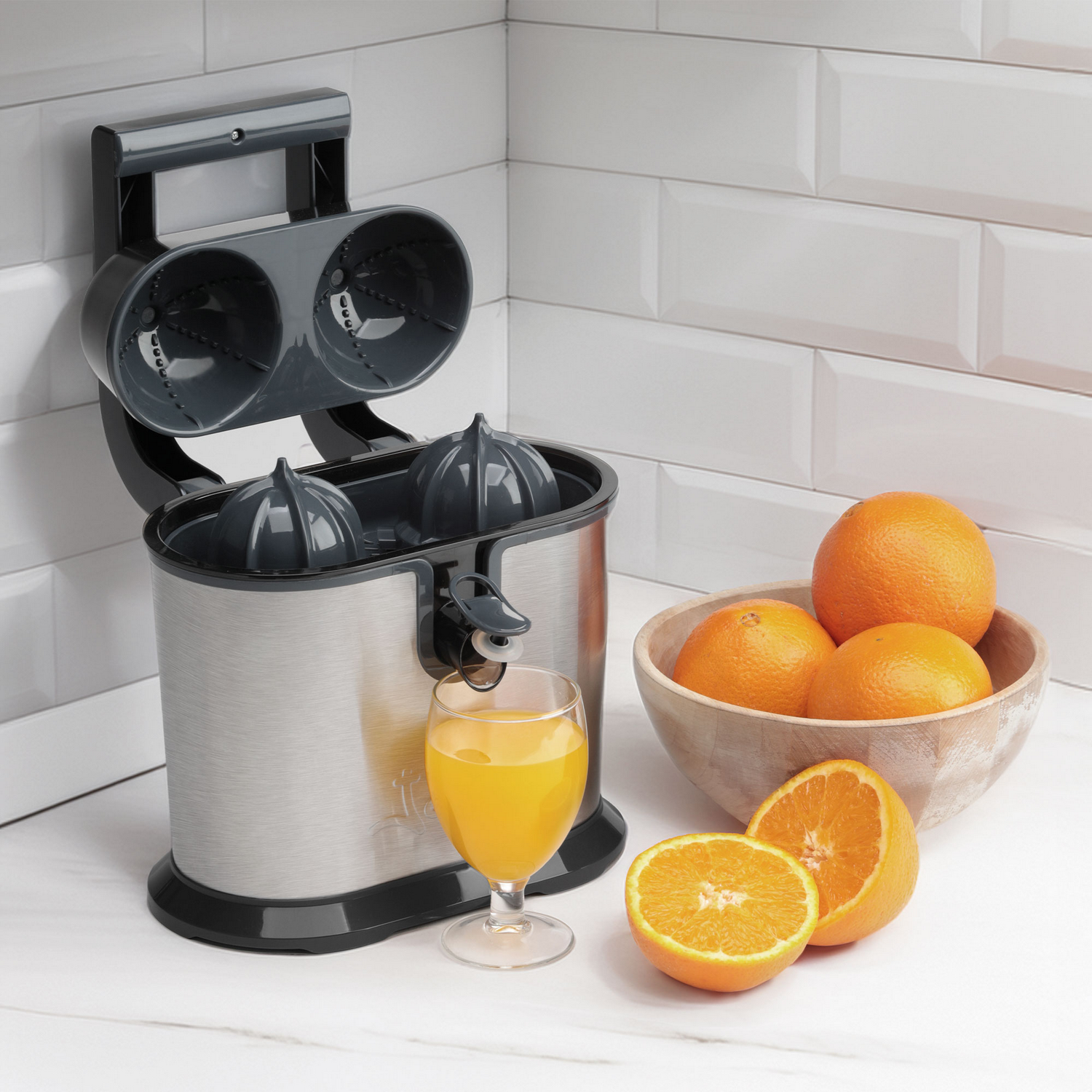 Exprimidor eléctrico con naranjas y un vaso de zumo. La escena está ambientada en una encimera blanca con fondo de azulejos blancos.