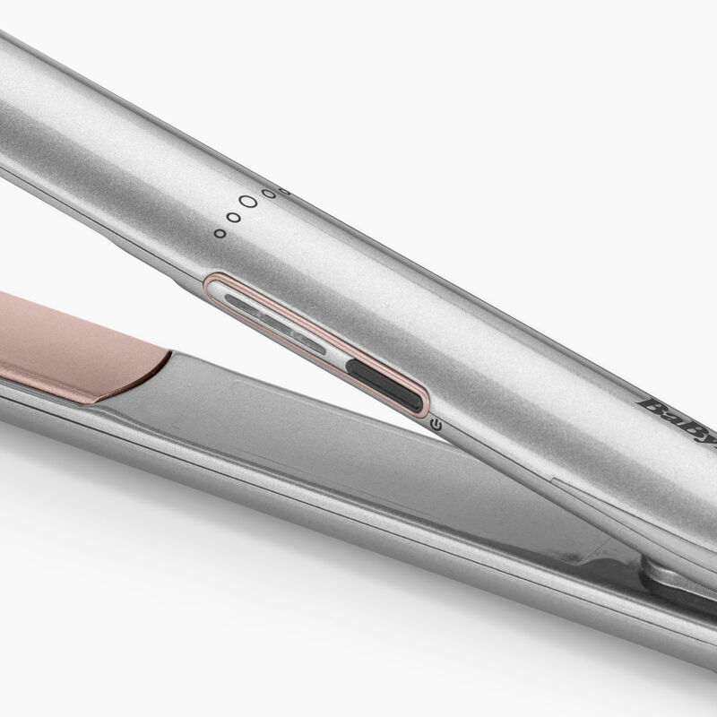 Primer plano de una plancha de pelo plateada con detalles en oro rosa. Botón de encendido visible. También es visible la marca 'BaByliss'.