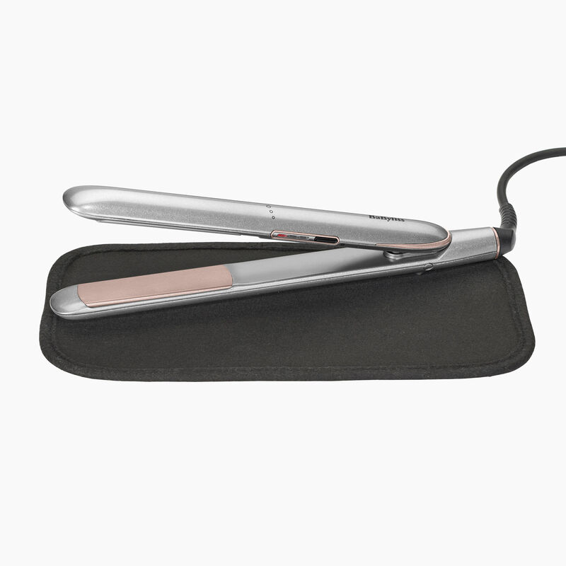 Plancha de pelo plateada con detalles en oro rosa sobre una alfombrilla negra resistente al calor, con cable de alimentación negro, sobre fondo blanco.