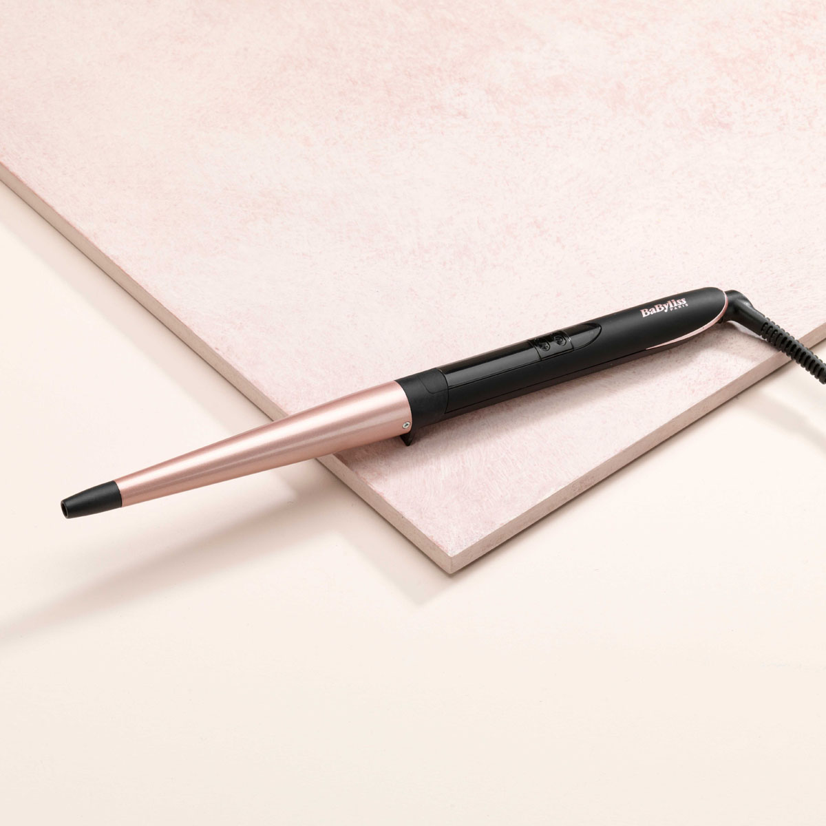 Una varilla rizadora con mango negro y barril oro rosa descansa sobre baldosas rosas. El cable es negro.