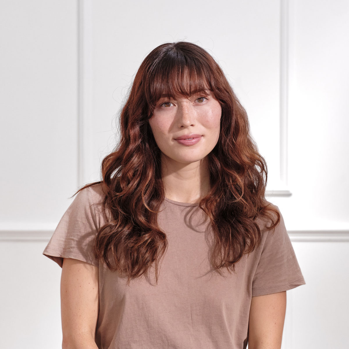 Una mujer con cabello rojo y una camisa marrón sonríe. Fondo blanco con molduras.