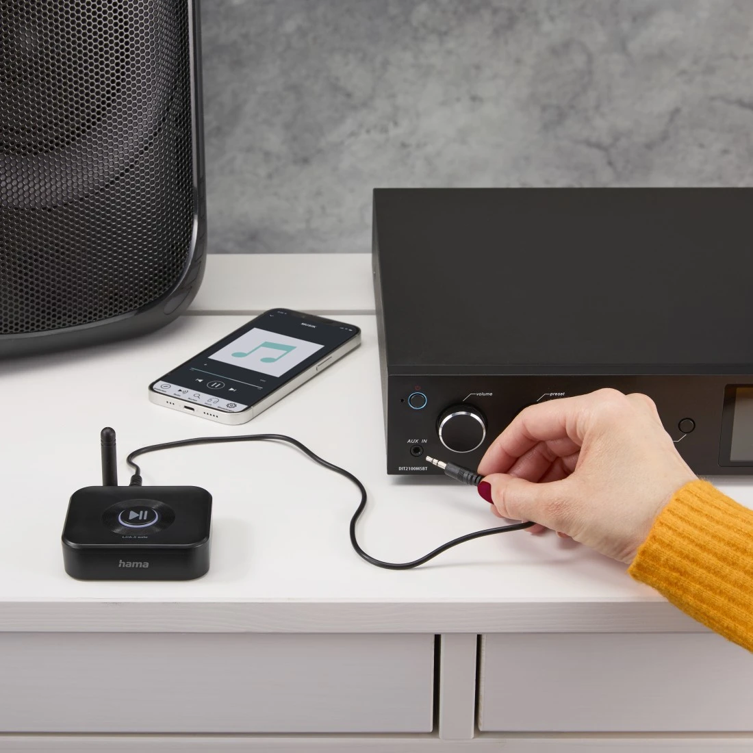 Una persona enchufa un conector de audio a un estéreo negro. Un teléfono muestra controles de música. Se ven un altavoz negro y un receptor de audio.