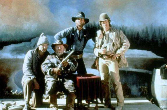 Vier Männer in Cowboy-Kleidung posieren. Einer hält ein Gewehr, ein anderer eine Pistole. Sie stehen vor einer gemalten Kulisse mit Fluss und Bäumen.
