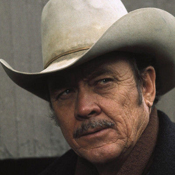 Nahaufnahme eines älteren Mannes mit Cowboyhut. Er hat einen Schnurrbart und einen ernsten Gesichtsausdruck. Der Hintergrund ist eine schlichte Wand.