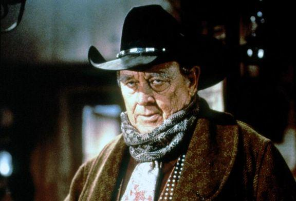 Ein älterer Mann mit Cowboyhut und Mantel blickt den Betrachter an. Er hat einen Schal und eine gemusterte Krawatte.