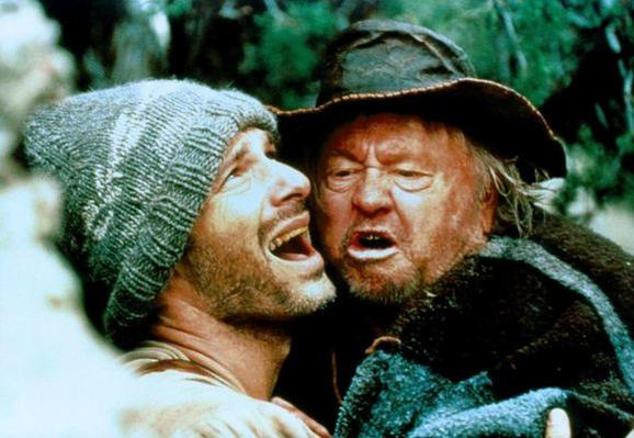 Zwei Männer umarmen sich und schreien, einer mit Strickmütze. Der ältere Mann trägt Hut und Mantel, Wald im Hintergrund.