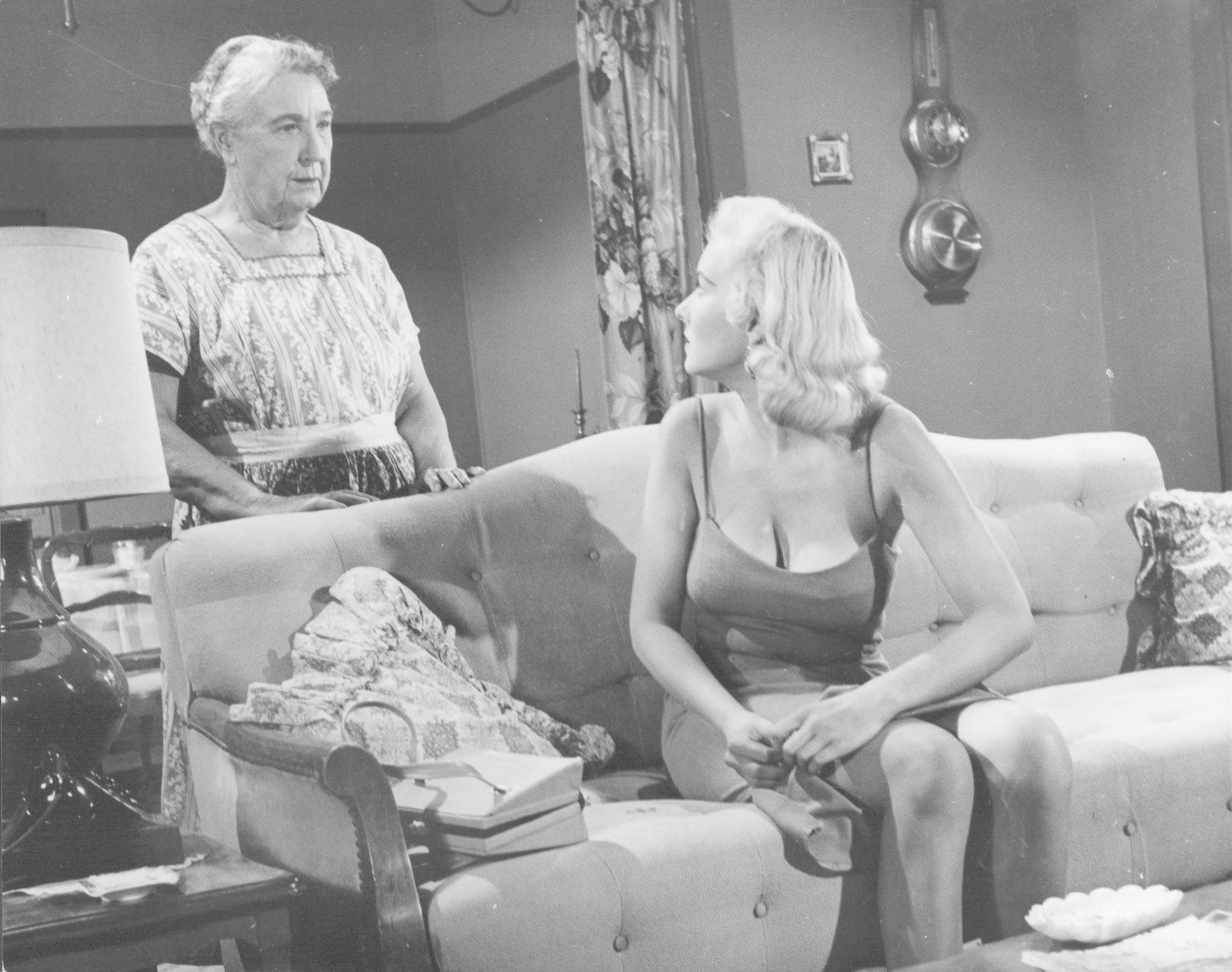 Zwei Frauen in einem Raum. Eine ältere steht, die andere, jünger, sitzt auf einem Sofa. Eine Lampe und eine dekorative Uhr sind sichtbar.