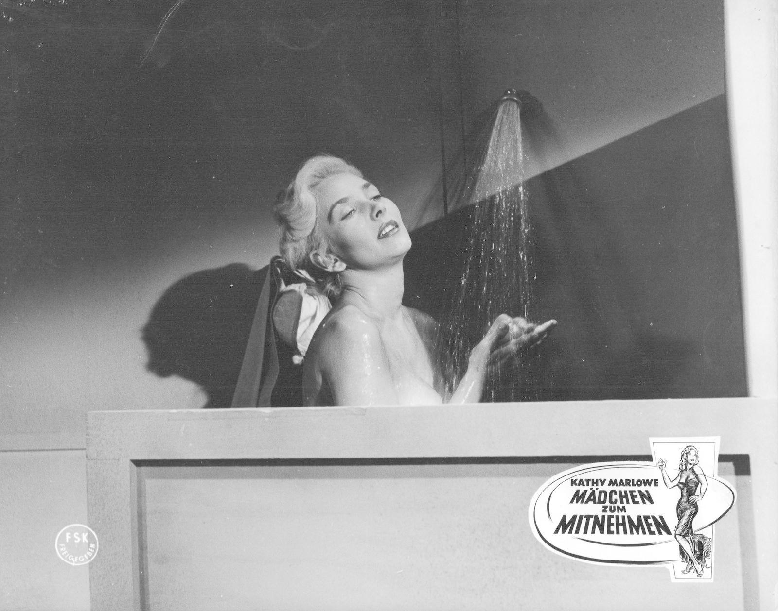 Eine Frau in der Dusche, die nach oben schaut. Wasser strömt auf sie herab. Ein Filmplakat ist im Hintergrund.