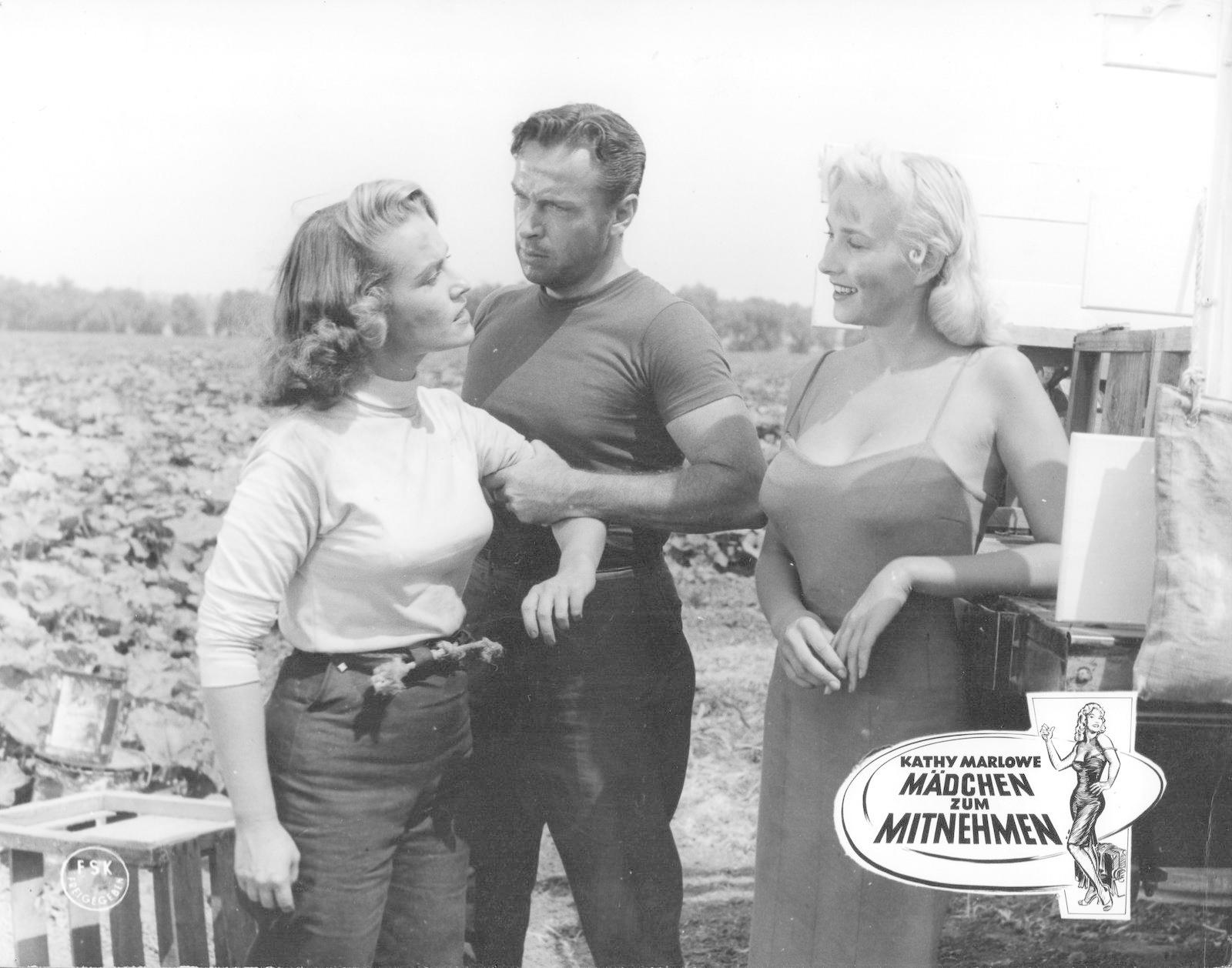 Drei Personen stehen auf einem Feld. Ein Mann im T-Shirt hält zwei Frauen. Ein Filmplakat mit deutschem Text ist rechts.
