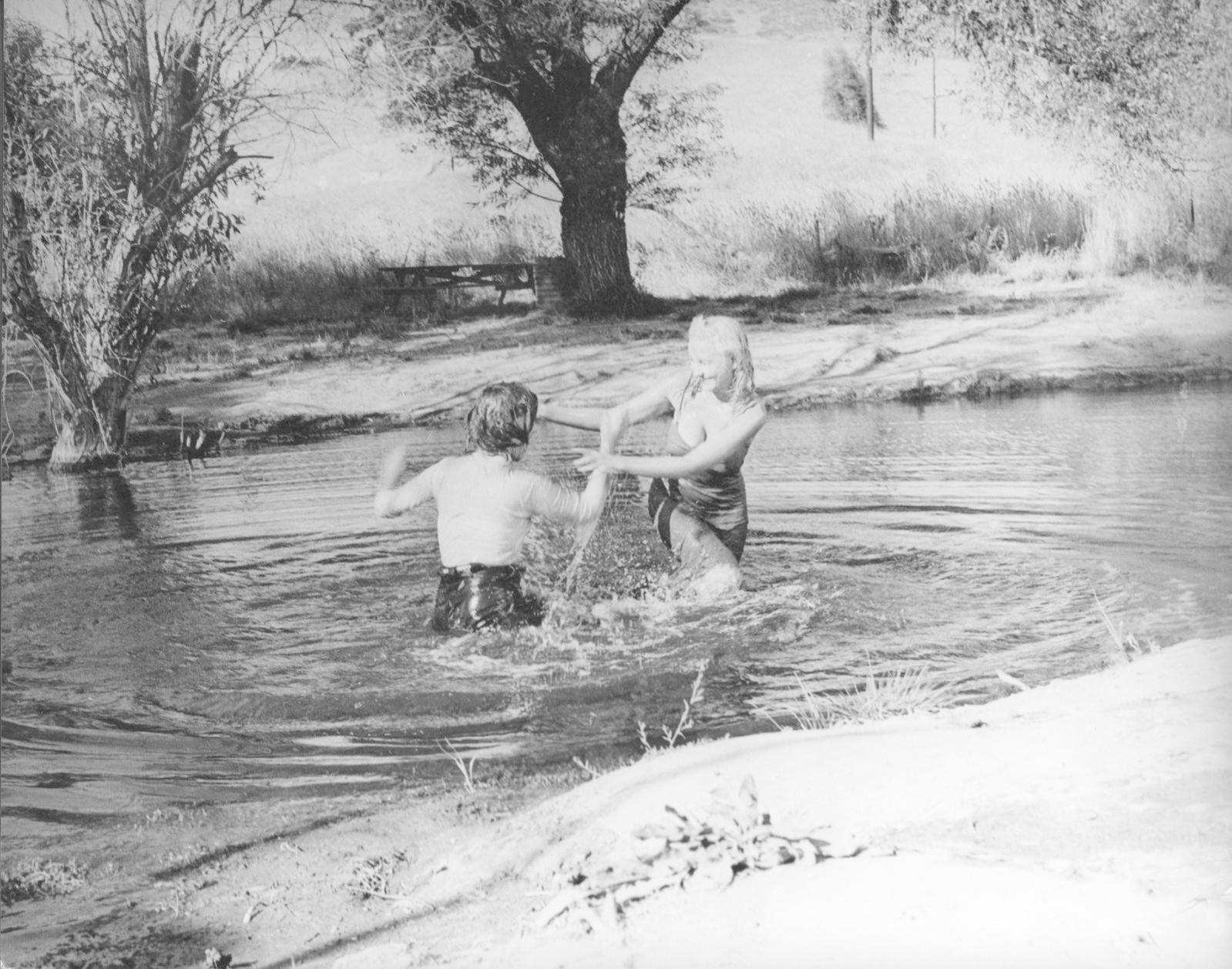Zwei Frauen planschen im Fluss. Eine trägt einen Badeanzug, die andere ein Hemd und eine Hose. Bäume und eine Picknickbank sind im Hintergrund.