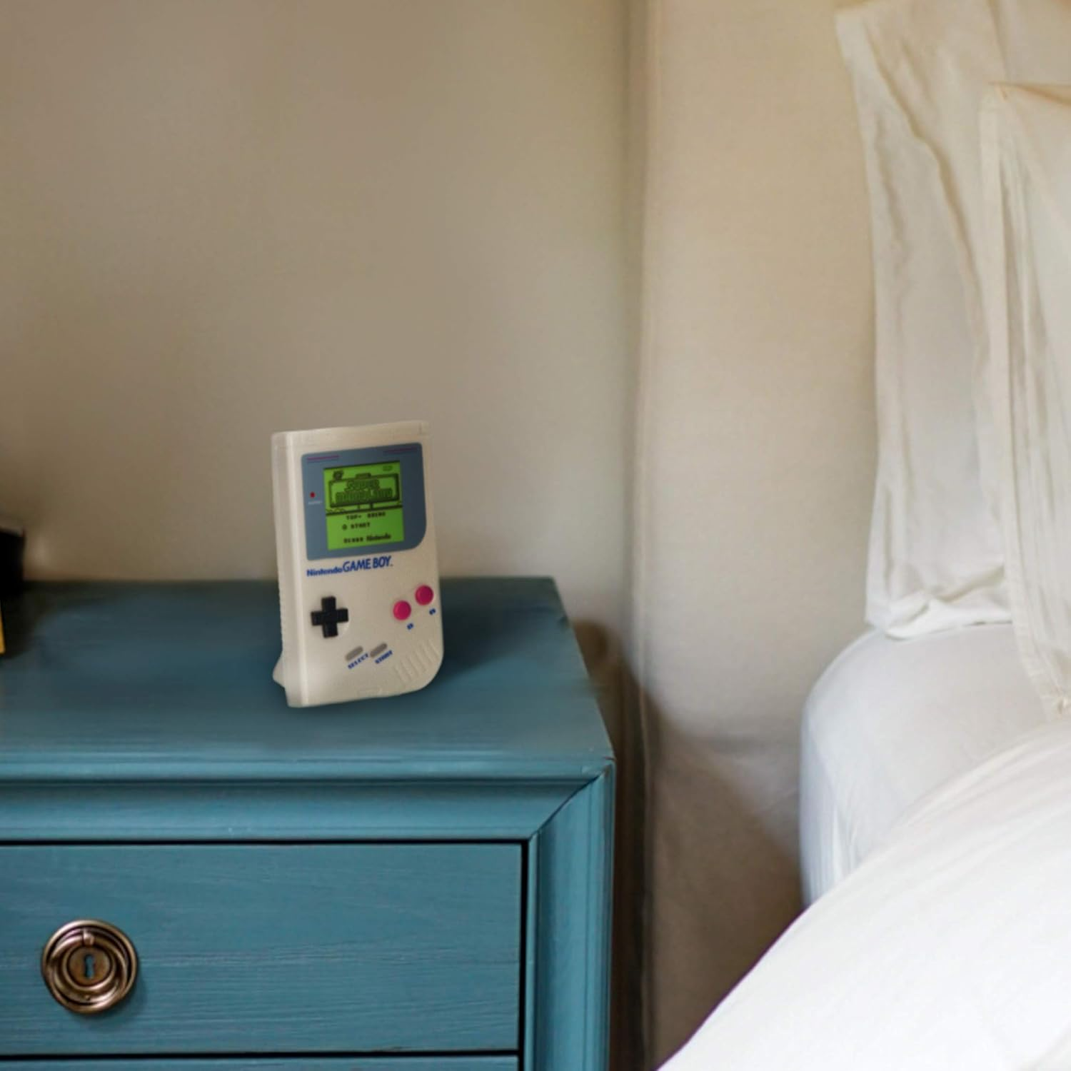 Una Nintendo Game Boy blanca está sobre una mesita de noche verde azulado, junto a una cama con ropa de cama blanca. Pared beige al fondo.