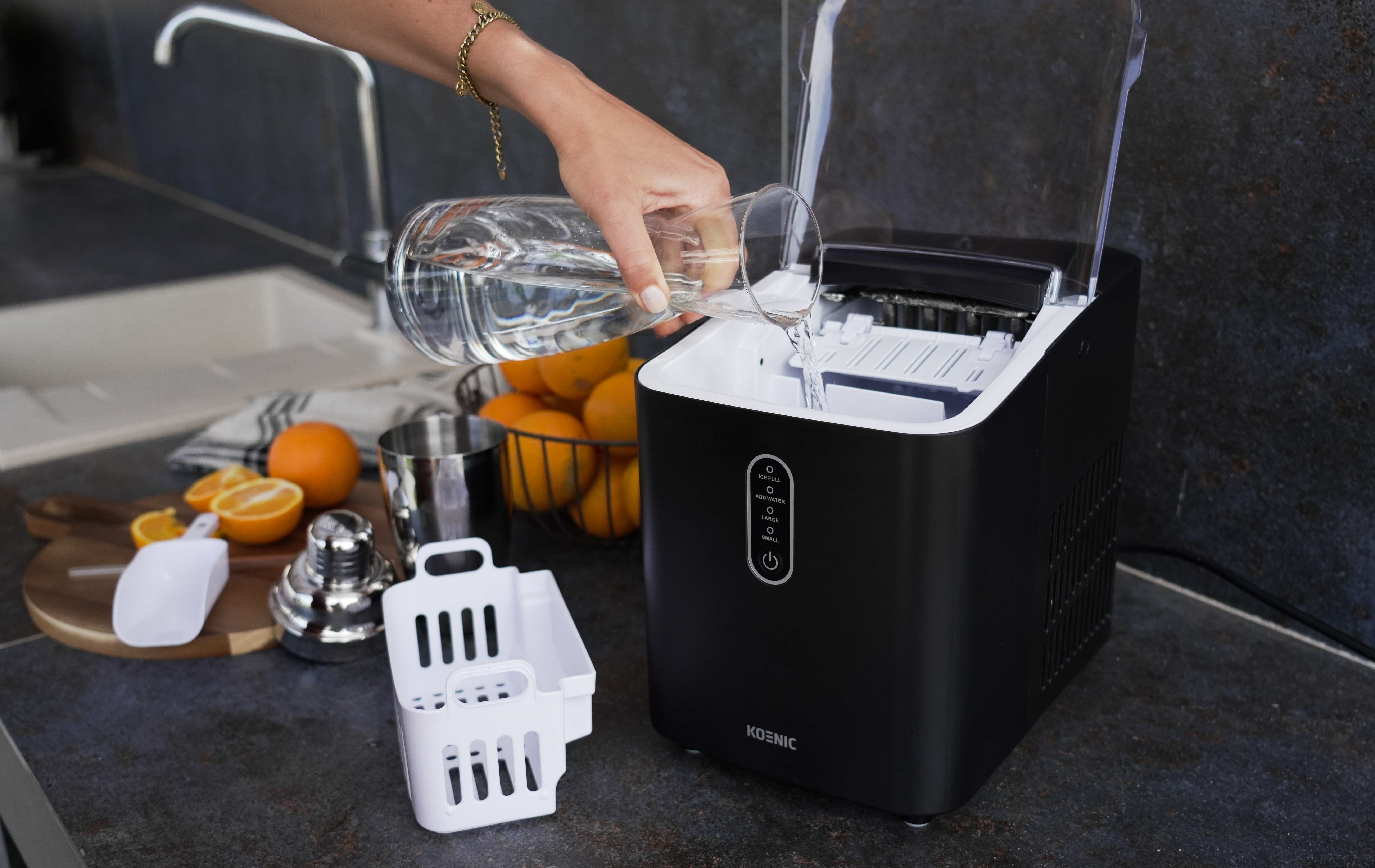 Ręka wlewa wodę ze szklanki do czarnej maszyny do lodu. W pobliżu znajdują się pomarańcze.