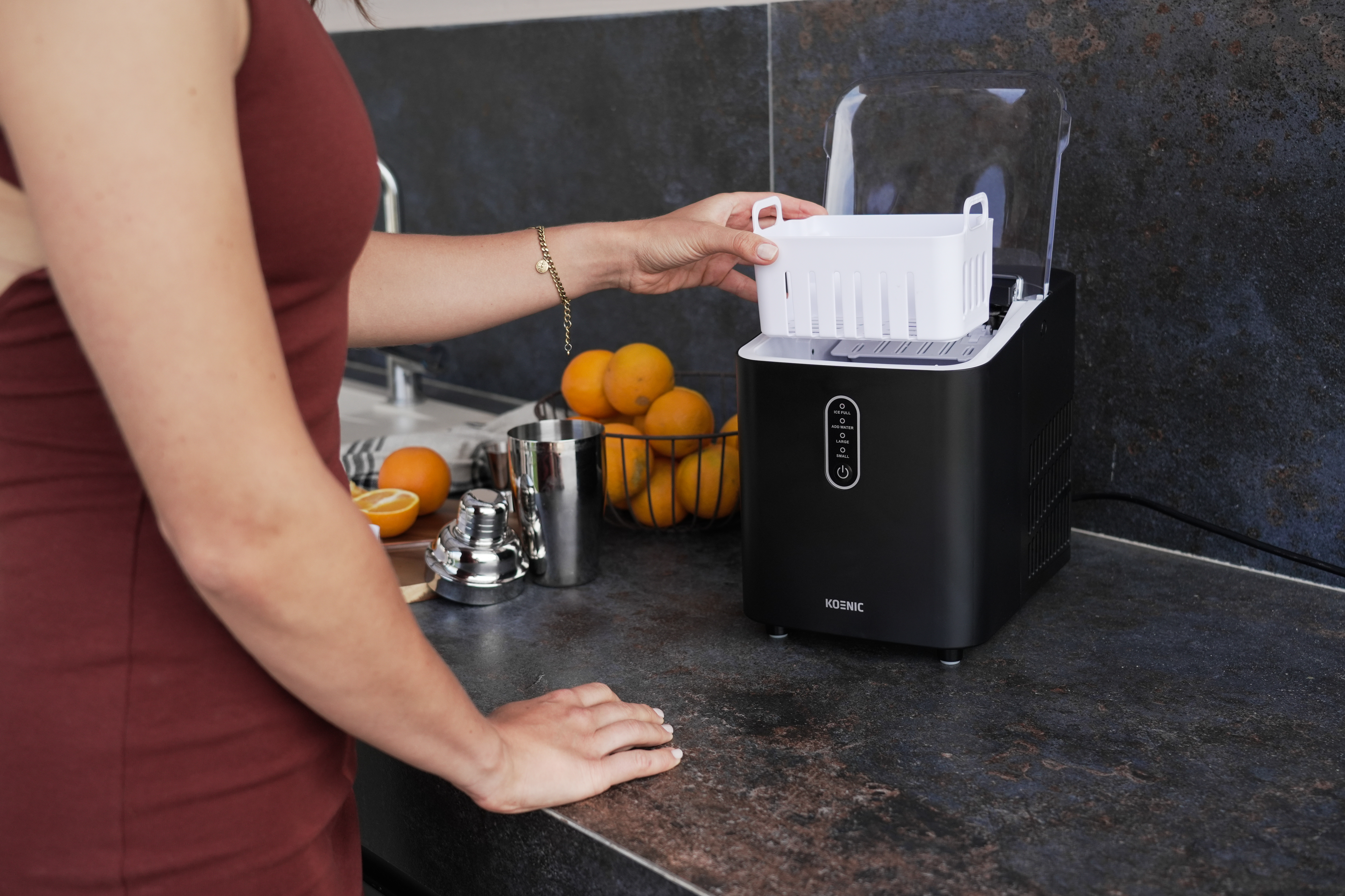 Kobieta wkłada biały pojemnik na lód do czarnej kostkarki na blacie kuchennym z pomarańczami.