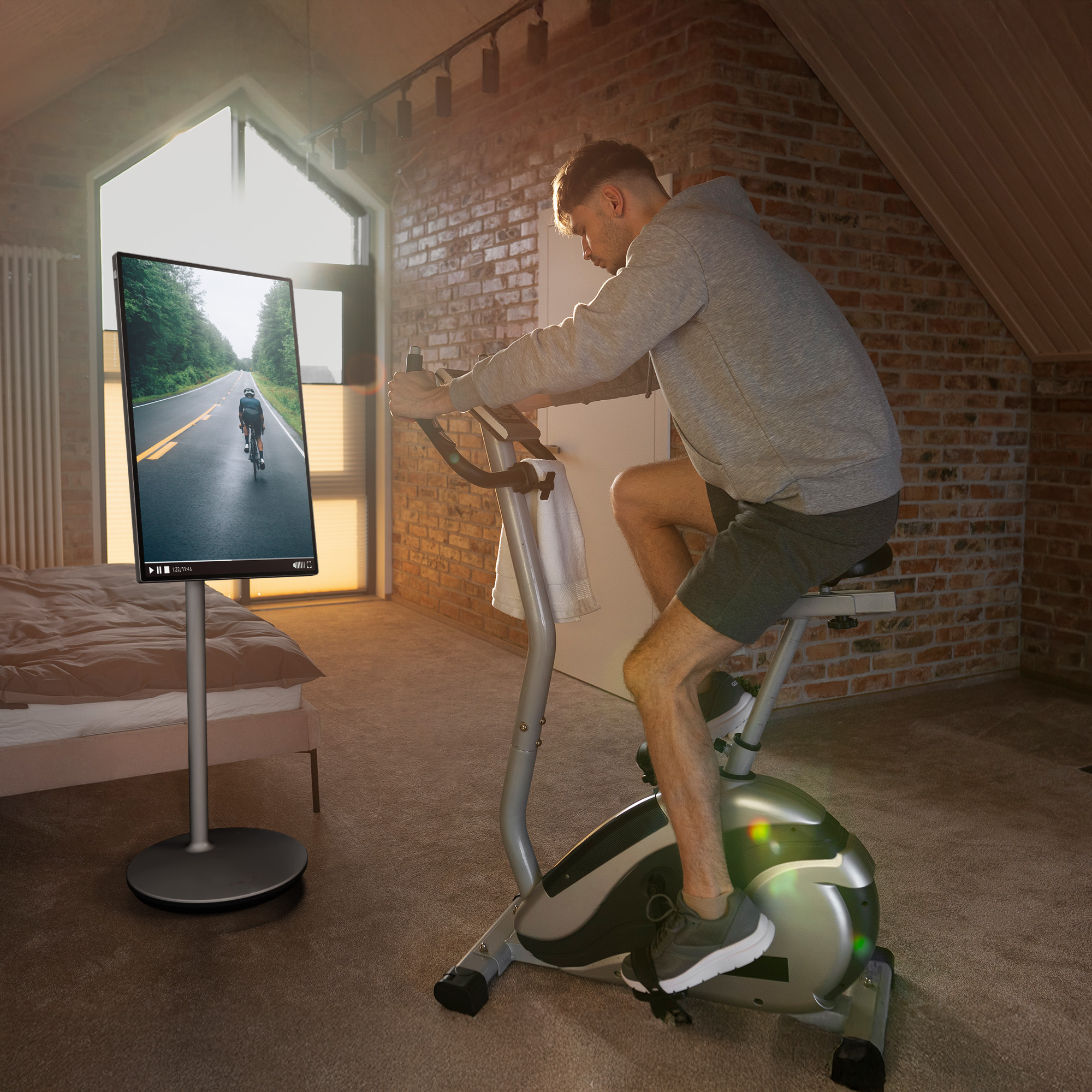 Hombre en bicicleta estática viendo un video de ciclismo en una pantalla. La habitación tiene pared de ladrillo y cama.