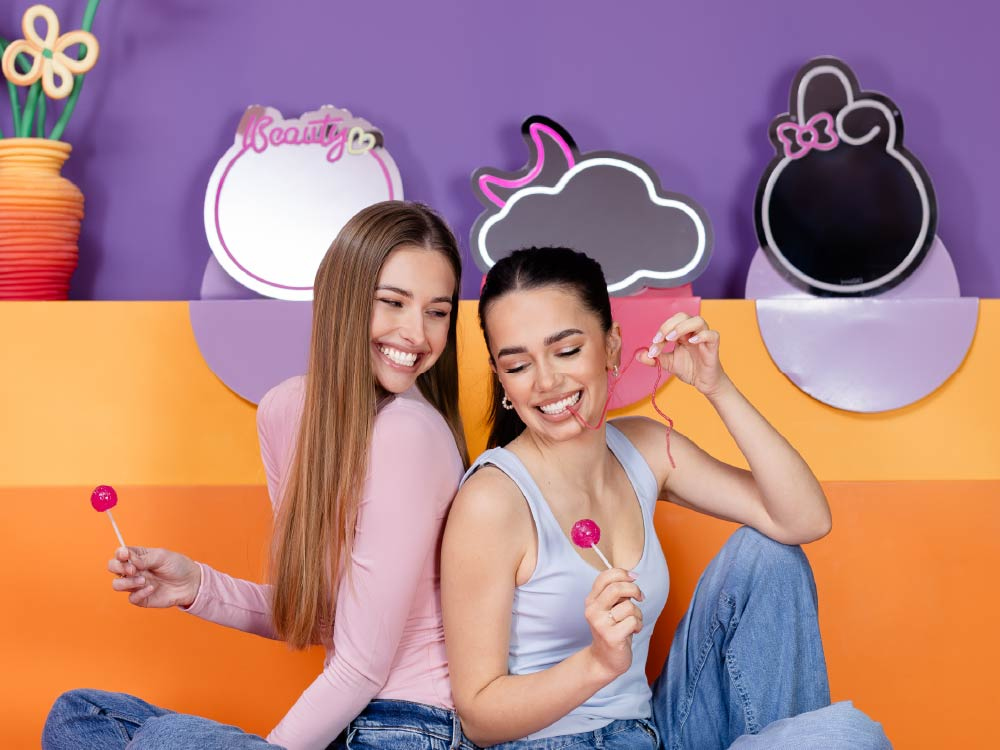 Dwie uśmiechnięte młode kobiety siedzą na pomarańczowej kanapie, jedna trzyma różowego lizaka, druga bawi się nitką.