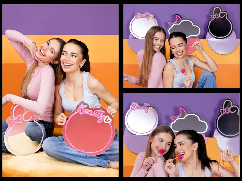 Dwie młode kobiety uśmiechają się, trzymając różowe lustra i lizaki na kolorowym tle.