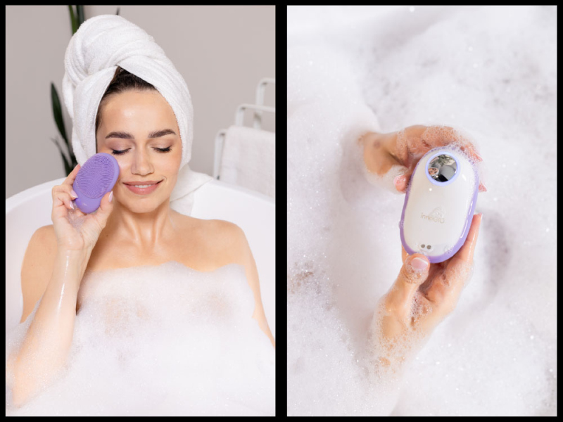 Kobieta z ręcznikiem na głowie używa fioletowej szczoteczki do mycia twarzy w kąpieli.