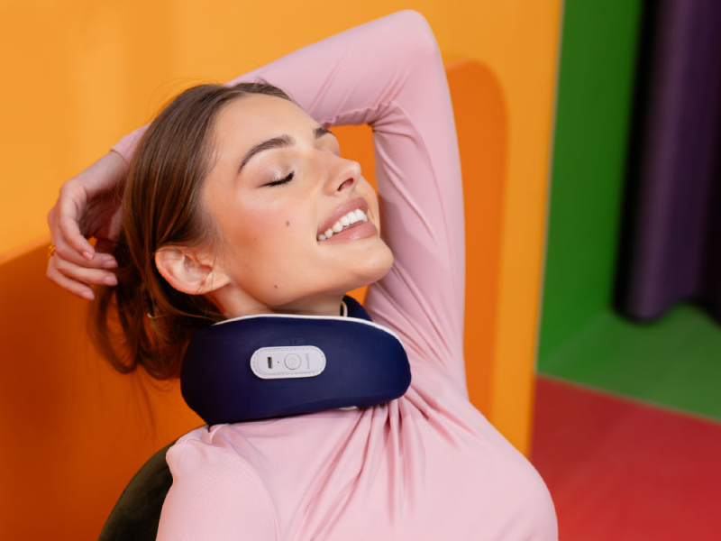 Kobieta z zamkniętymi oczami nosząca masażer karku i różowy top, kolorowe tło.