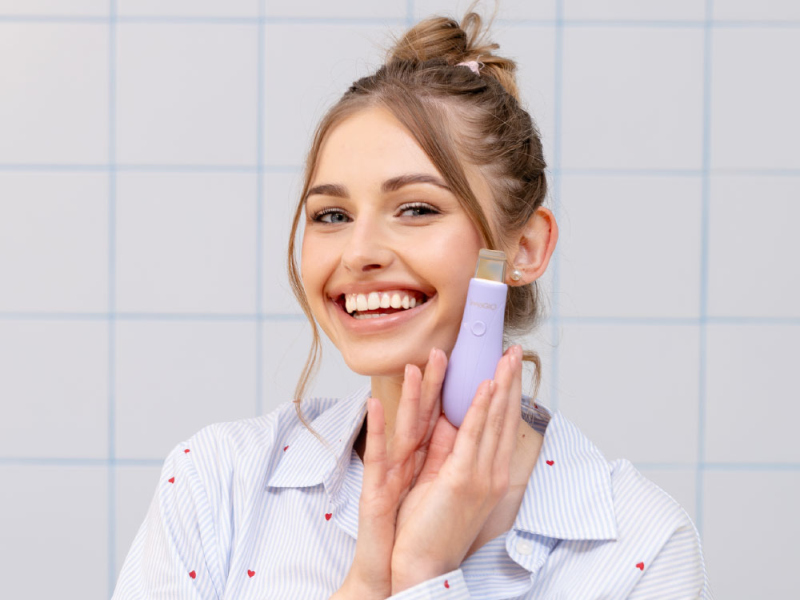 Uśmiechnięta kobieta w pasiastym T-shircie trzyma fioletowe urządzenie do oczyszczania twarzy. Tło w kafelkach.