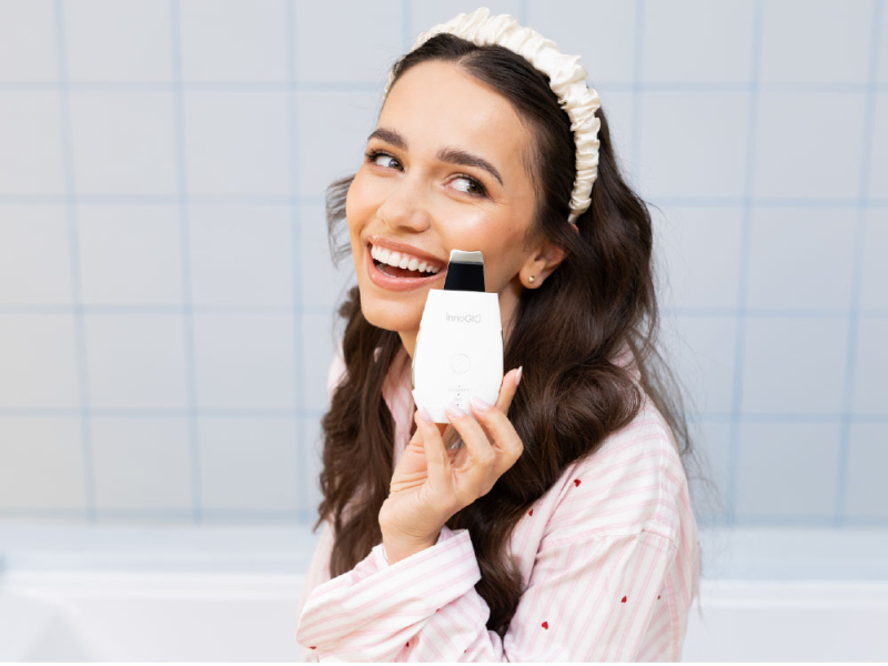 Uśmiechnięta kobieta w piżamie trzyma białe urządzenie do oczyszczania twarzy z czarną końcówką.