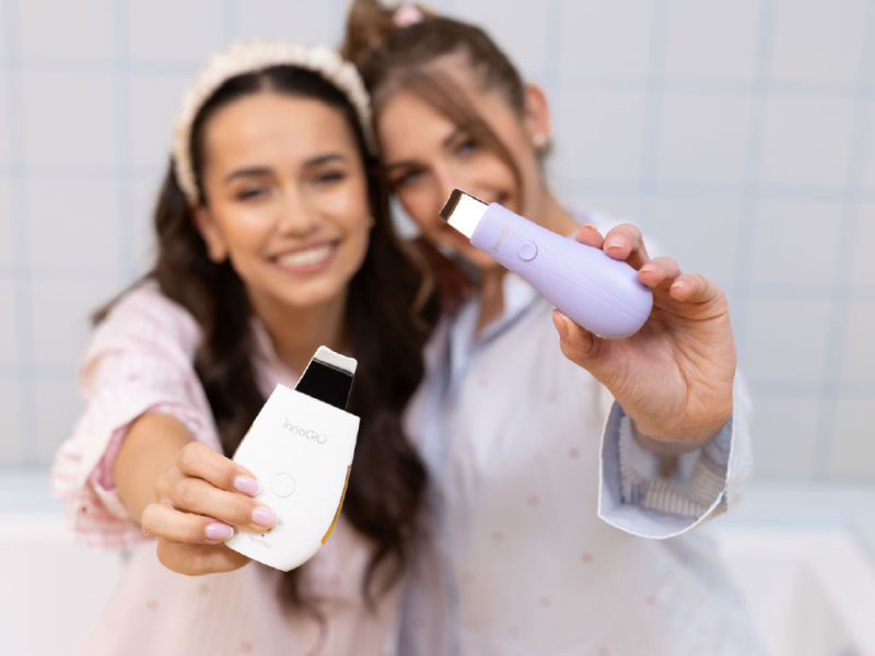 Dwie uśmiechnięte kobiety trzymają urządzenia do pielęgnacji skóry, białe i fioletowe.