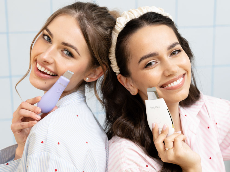 Dwie uśmiechnięte kobiety trzymające urządzenia do oczyszczania twarzy. Jedna trzyma lawendowe, druga białe urządzenie.