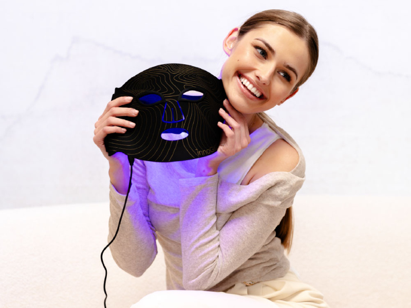 Uśmiechnięta kobieta trzymająca czarną maskę LED z złotymi liniami i niebieskimi światłami.