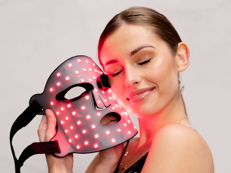 Kobieta trzyma maskę na twarz z czerwonymi diodami LED blisko twarzy, światło pada na jej policzek.
