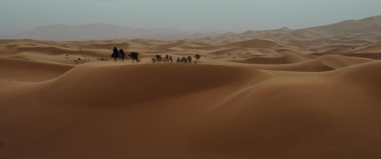 Eine Karawane aus Menschen und Kamelen durchquert eine riesige Wüste. Sanddünen dominieren die Landschaft.