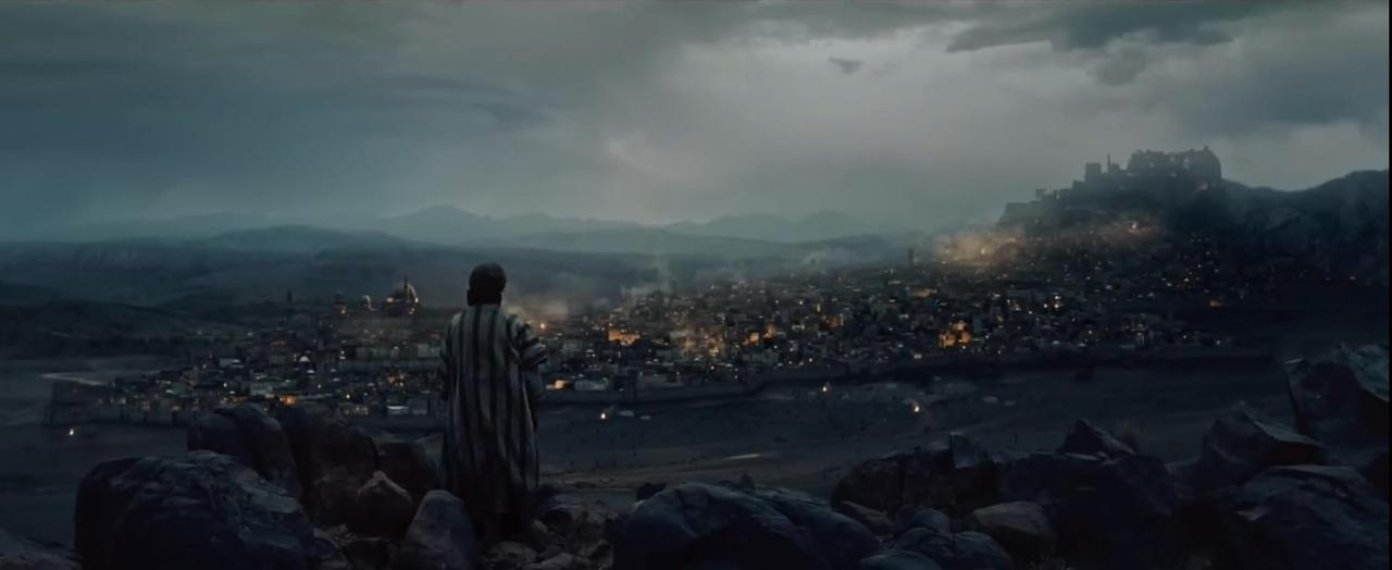 Eine Person in gestreiften Gewändern blickt auf eine Stadt, die nachts beleuchtet ist. Berge und dunkle Wolken im Hintergrund.