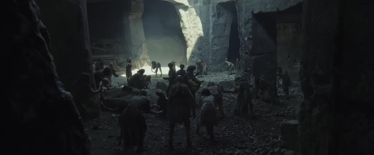 In einer dunklen Höhle versammelt sich eine Gruppe von Menschen. Einige stehen, andere sitzen oder bücken sich. Die Szene wirkt düster.