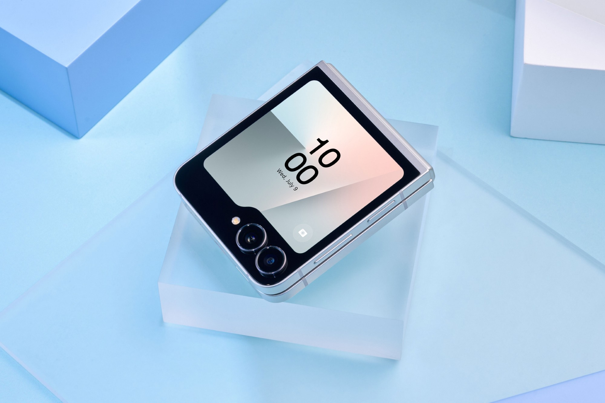 Srebrny składany telefon na półprzezroczystym bloku, otoczony niebieskimi kształtami geometrycznymi. Telefon wyświetla czas. Miękkie oświetlenie.