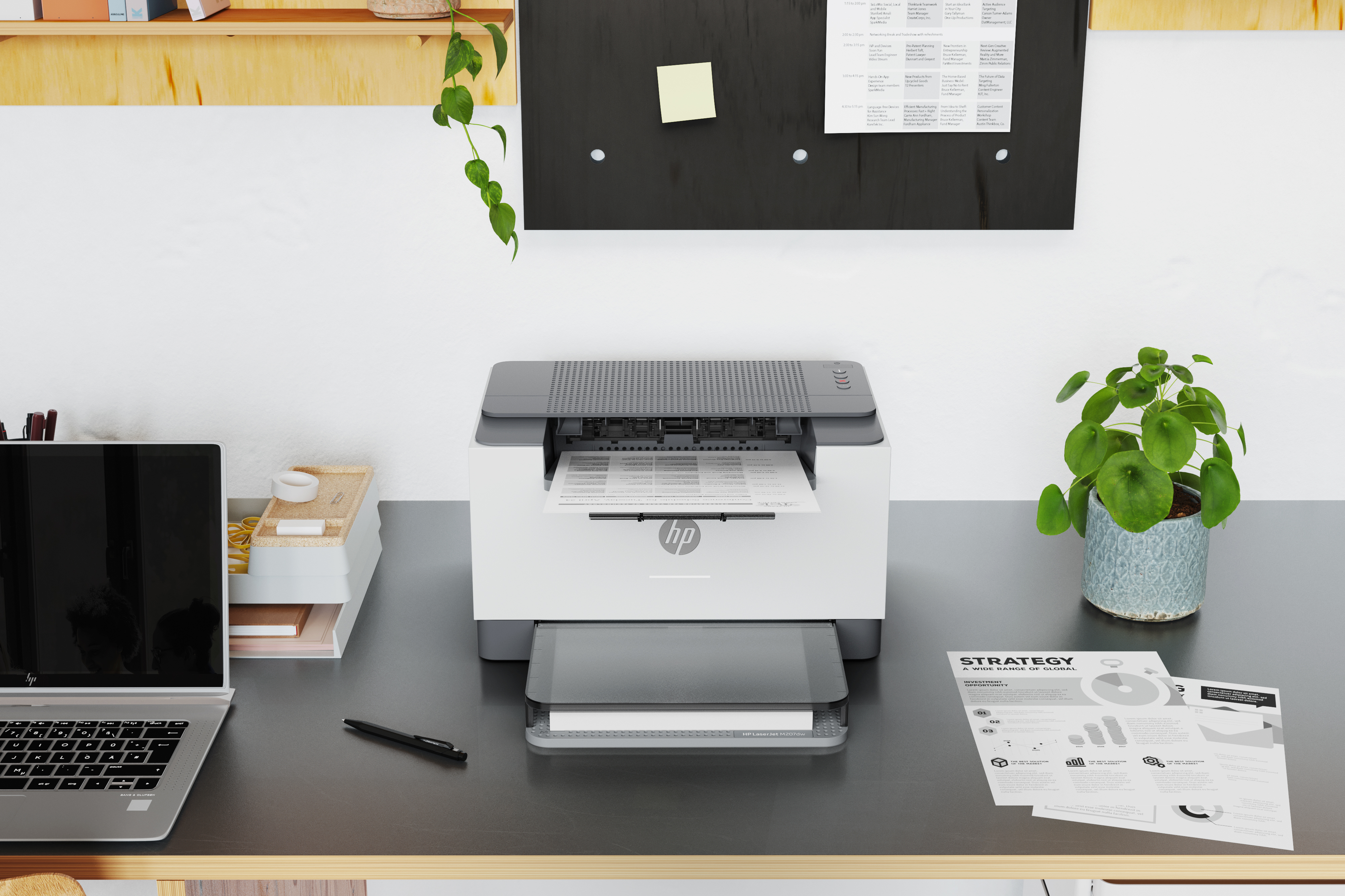 Een bureau met een printer, laptop en documenten. Een plant hangt van bovenaf.